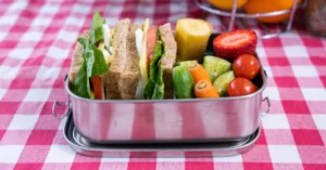 Metal lunchbox with a sandwich, fruit and vegetables on red and white chequered tablecloth