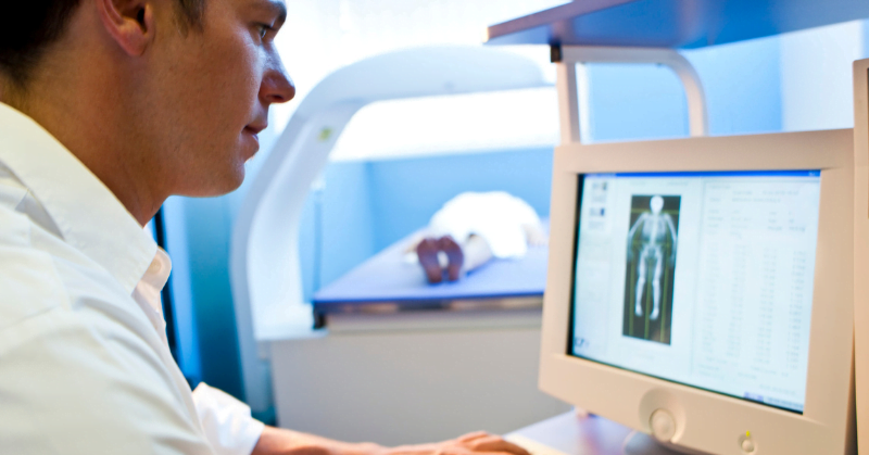 Clinician viewing scan image on computer with person laying down in bone density scan machine in the background