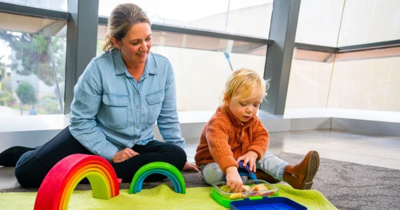 Mother and child sitting on the floor with rainbow blocks while the child eats from a lunchbox