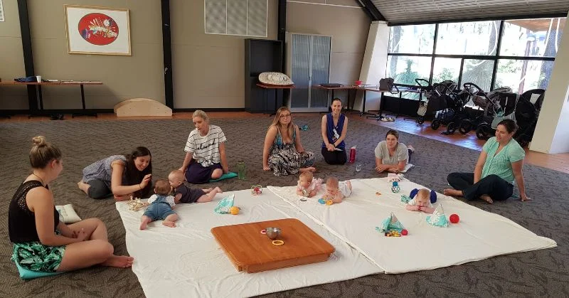 Mothers sitting around the edge of a white mat that has babies laying on it 