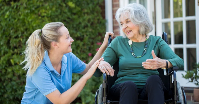 Older lady in wheelchair outside in a garden while younger female carer crouches on the ground talking to each other.