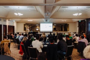 A conference room filled with attendees seated at round tables, attentively listening to a presentation. At the front of the room, Professor Gary Frost stands beside a podium, speaking while a large projector screen displays slides.