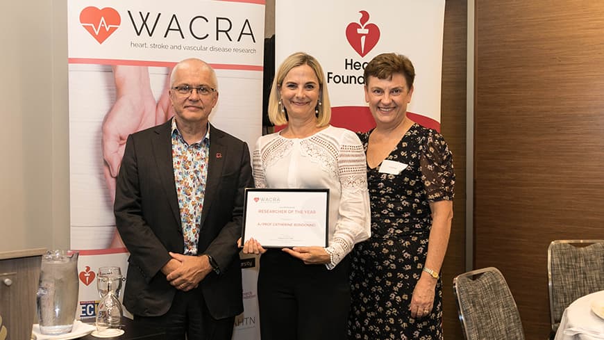 Associate Professor Catherine Bondonno holds her framed award with two academics, WACRA pull-up banners are placed behind them.