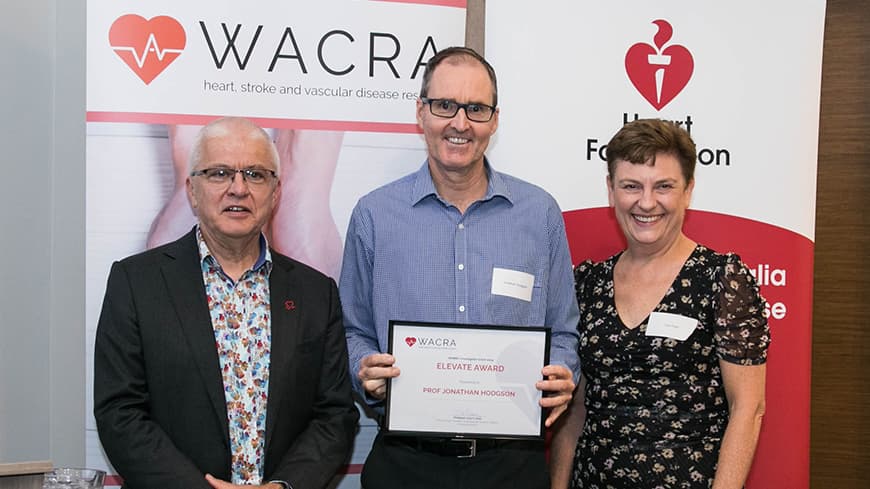 Professor Jonathan Hodgson holds his framed award with two academics, WACRA pull-up banners are placed behind them.