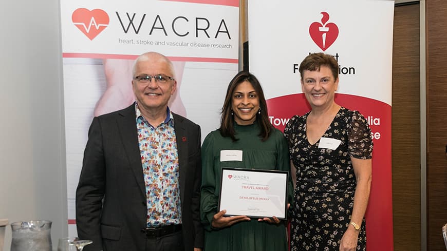 Dr Nilufeur McKay holds her framed award with two academics, WACRA pull-up banners are placed behind them.